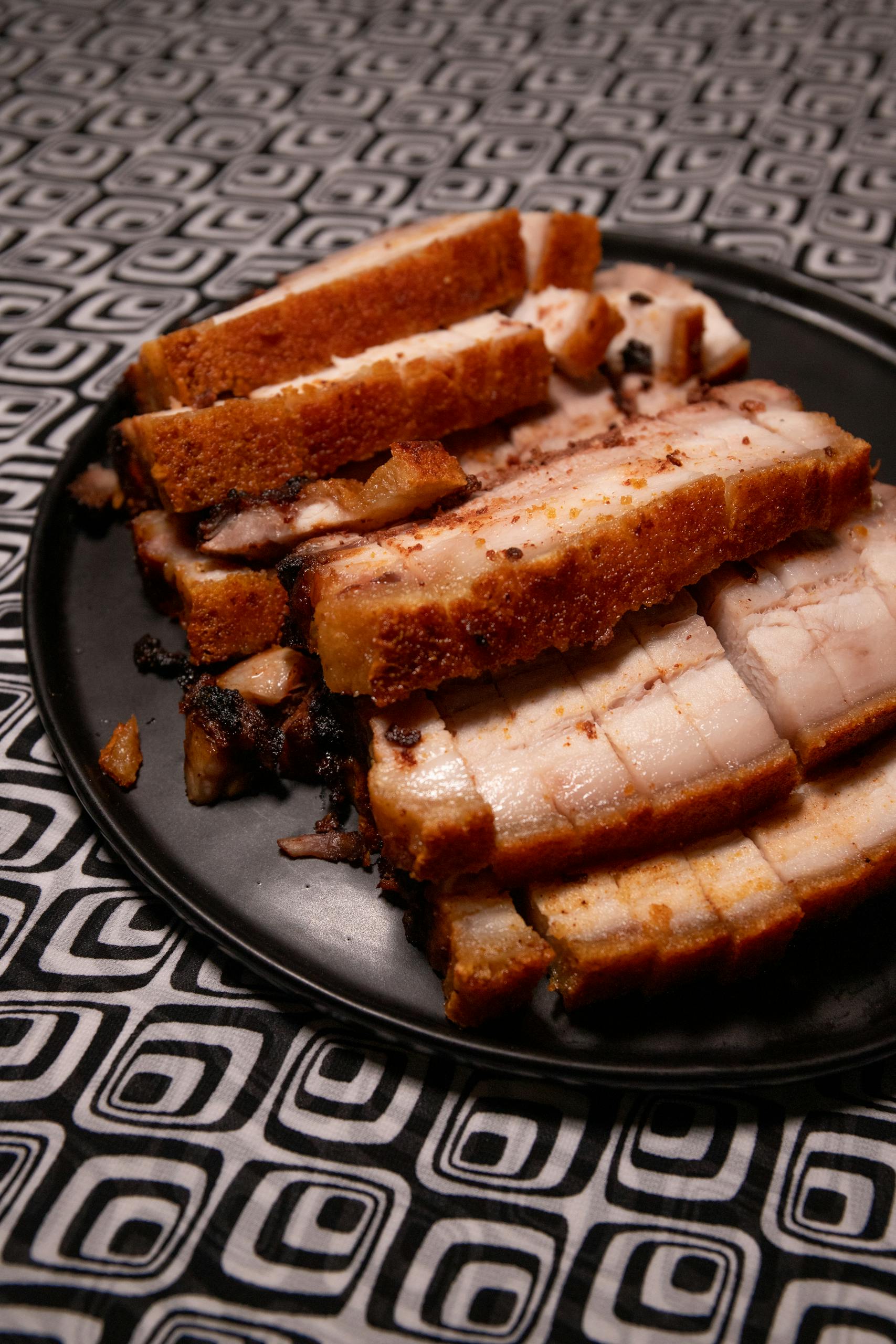Delicious sliced crispy pork belly served on a black plate against a patterned tablecloth.